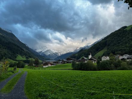 Ein Tal mit grünen Wiesen, ein paar Häuser und im Hintergrund Berge mit etwas Schnee. Der Himmel ist wolkenverhangen.