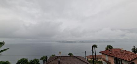 Im Vordergrund ein paar Hausdächer, dahinder der See, in der ferne flaches Land. Darüber ein grauer, Wolkenverhangener Himmel.