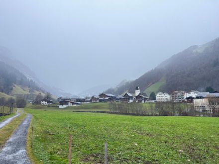 Zu sehen ist graue Bewölkung über dem Dorf Elm, im Vordergrund ein nasser Feldweg und eine Wiese. 