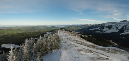 Leicht verschneite Berge und Bäume, dahinter ein meist blauer Himmel.
