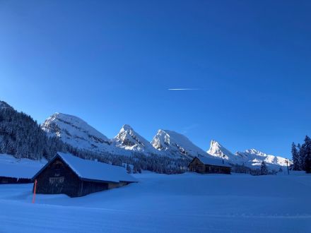 Eine verschneite Landschaft mit Hütten, Loipe und Berggipel im Hintergrund.