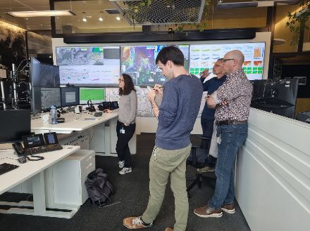 Vier Leute die auf mehrere Bildschrime schauen. Im Hintergrund sieht man eine Wand mit grossen Monitoren und vielen Wetterinformationen.