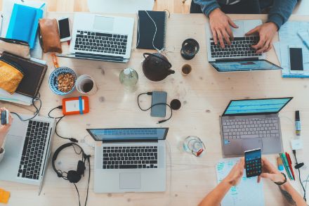 Das Bild zeigt einen grossen Holztisch mit mehreren Laptops, Smartphones und Arbeitsmaterialien, während Personen daran arbeiten und interagieren.