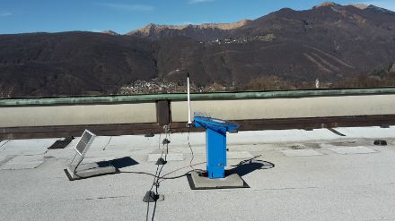 Ein blauer Hagelsensor aus Metall mit einer Polycarbonat Messcheibe darauf, daneben ein Solarmodul.