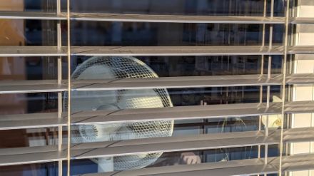 Blick von aussen durch ein Fenster mit halb gekippten Storen. Im Zimmer steht ein Ventilator am Fenster.