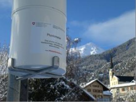 Im Vordergrund ein Pluviometer, im Hintergrund schneebedeckte Wälder und Berge.
