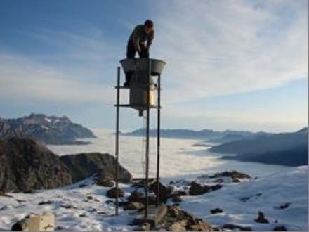 Im Bild eine Person beim Leeren des Totalisators, im Hintergrund Berggipfel, die aus dem Nebelmeer ragen