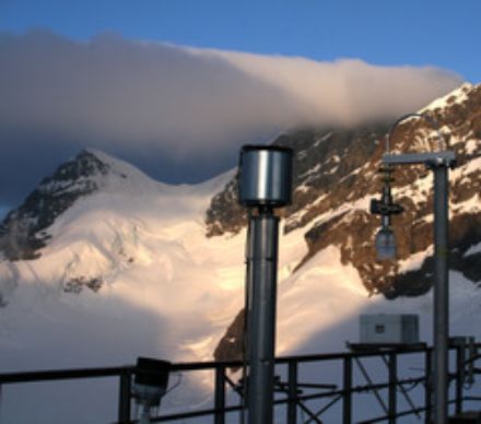 Die Inlets stehen auf einer Terrasse im Freien. Das Inlet in der Bildmitte besteht aus einem vertikal angebrachten Rohr mit einem Metallbehälter an der Spitze, durch den die Luft angesaugt wird. Im Hintergrund Berge und Gletscher.