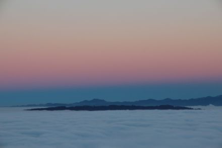 Blick über ein Nebelmeer zu den Bergen. Dahinter steigt ein schmaler, dunkler Saum - der Erdschatten - in die Höhe. Scharf abgetrennt sind darüber rötliche Dämmerungsfarben zu sehen.