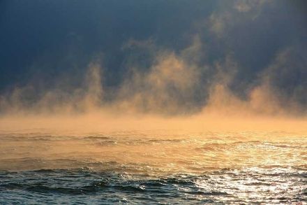 Verdunstungsnebel über einem See, der im Sonnenlicht orange leuchtet.
