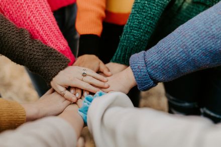 Mehrere Hände liegen übereinander als Zeichen von Zusammenhalt. Die Personen tragen bunte Strickpullover, einige Hände sind mit Ringen geschmückt. Das Bild strahlt Gemeinschaft und Teamgeist aus.