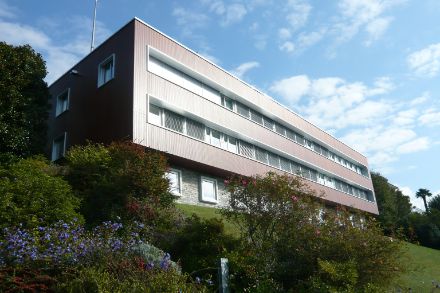 Modernes, mehrstöckiges Gebäude an einem Hang mit grossen Fenstern, umgeben von blühenden Sträuchern und blauem Himmel im Hintergrund.